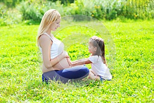 Happy pregnant woman, mother and little daughter child sitting on grass in summer