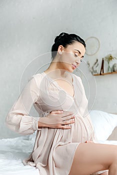 Happy pregnant woman in nightie touching tummy while sitting on bed