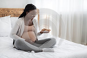 Happy pregnant woman with plate of cookies sitting in bed at home