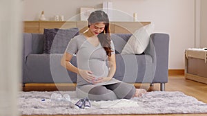 Happy Pregnant Woman sit on carpet holding stroking her big belly with diaper,feeding bottle,baby shoes at home.Pregnancy of young
