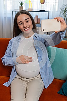 Happy pregnant woman sits on sofa taking selfie using smartphone touching belly enjoying motherhood