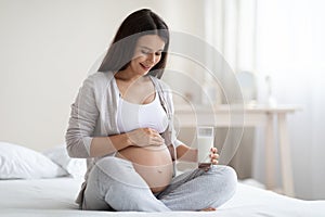 Happy pregnant woman sitting on bed, holding glass of milk