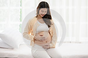 Happy Pregnant Woman sitting on bed holding and stroking her big belly at home,Pregnancy of young woman enjoying with future life