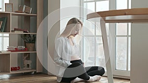 Happy pregnant woman is sitting on the floor of room near large window