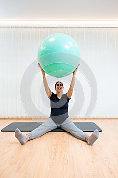 A happy pregnant woman sitting on a mat doing pilates exercises with a ball