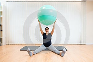 A happy pregnant woman sitting on a mat doing pilates exercises with a ball