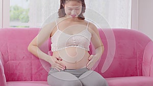 Happy Pregnant Woman sitting on pink couch holding and stroking her big belly at cozy home,Pregnancy of young woman enjoying with