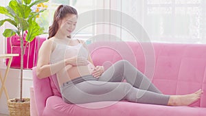 Happy Pregnant Woman sitting on pink couch holding and stroking her big belly at cozy home,Pregnancy of young woman enjoying with