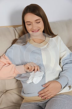 Happy pregnant woman sitting on sofa with baby socks indoors