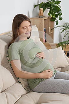 Happy pregnant woman sitting on sofa and touching her belly in living room