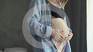 Happy pregnant woman standing on bedroom holding and stroking her big belly at home, close up, pregnancy of young woman