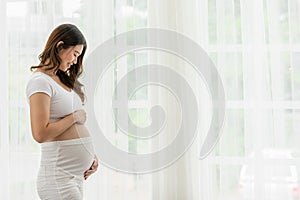 Happy Pregnant Woman standing in front of windows and stroking her big belly with love at cozy home,Pregnancy of young woman enjoy