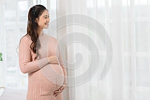 Happy Pregnant Woman standing in front of windows and stroking her big belly with love at cozy home,Pregnancy of young woman enjoy