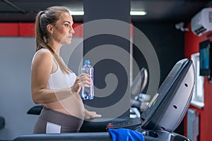 Happy pregnant woman is tired of running on a treadmill and drinking water from a plastic bottle in the gym looking at