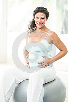 Happy pregnant woman touching her belly while sitting on exercise ball