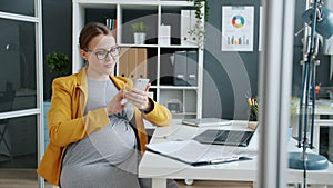 Happy pregnant woman using smartphone enjoying online content working at desk in office