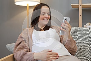 Happy pregnant woman using smartphone at home browsing pregnancy app while sitting on couch in living room smiling expectant lady