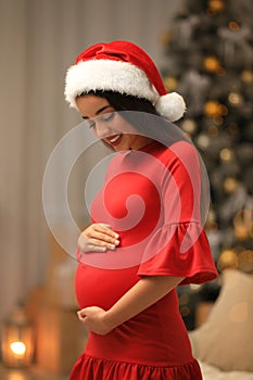 Happy pregnant woman wearing Santa hat in room for Christmas. Expecting baby