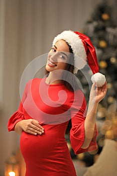 Happy pregnant woman wearing Santa hat in room for Christmas. Expecting baby