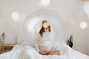 Happy pregnant woman in white holding belly bump and relaxing on white bed at home. Stylish pregnant mom hugging belly with love