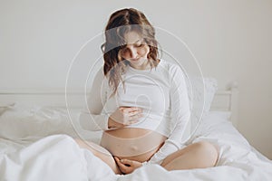 Happy pregnant woman in white holding belly bump and relaxing on white bed at home. Stylish pregnant mom hugging belly with love