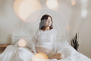 Happy pregnant woman in white holding belly bump and relaxing on white bed at home. Stylish pregnant mom hugging belly with love