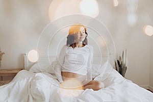 Happy pregnant woman in white holding belly bump and relaxing on white bed at home. Stylish pregnant mom waiting for baby.