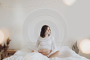 Happy pregnant woman in white holding belly bump and relaxing on white bed at home. Stylish pregnant mom waiting for baby.