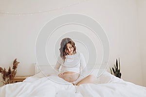 Happy pregnant woman in white holding belly bump and relaxing on white bed at home. Stylish pregnant mom waiting for baby.