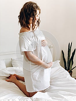 Happy pregnant woman in white holding belly bump and relaxing on white bed at home. Stylish pregnant mom waiting for baby.