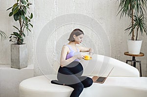 Happy pregnant young adult woman sitting on a sofa, working on a laptop