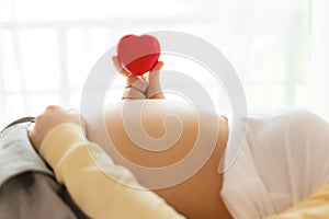 Happy pregnant young woman holding red heart in hand on top of mother belly in hospital bed. prenatal, pregnancy, motherhood,