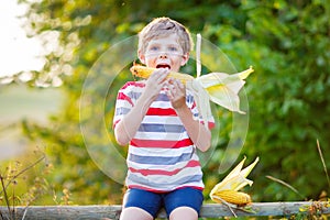 Happy preschool kid boy with glasses holding ears of corn on farm in field, outdoors. Funny child hild having fun with