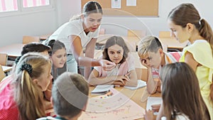 Happy preteen children and female teacher playing together educational board game in classroom at elementary school