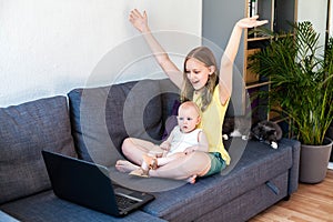 Happy preteen girl with her infant sister looking at laptop screen and waving hands while having video call.