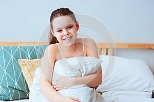 happy preteen girl sitting on bed and hugs cozy pillow