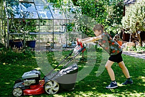 Happy preteen kid boy with lawn mower. Portrait of smiling teenager child working in garden, trimming grass. Garden