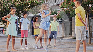 Happy preteen kids of different nationalities skipping on chinese jump rope in courtyard on summer day