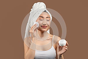 Happy pretty asian woman cleansing her skin with cotton pad, holding jar with cream and smiling over brown background