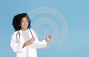 Happy pretty black millennial lady doctor in white coat point fingers up to free space, recommends ad and offer