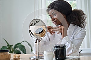 Happy pretty black young woman looking in mirror doing makeup.