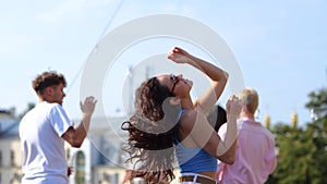 Happy pretty female in sunglasses dancing on the roof open air party while moving hands up, friends on the background