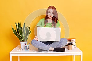 Happy pretty ginger woman sitting on table and working with laptop computer, looks with concentrated facial expression at device