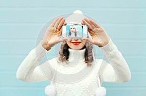 Happy pretty girl taking photo self portrait on smartphone over blue background in a winter knitted clothes