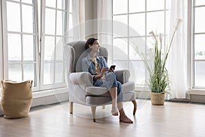 Happy pretty middle aged woman holding cellphone, sitting in armchair