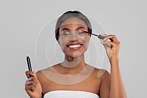 Happy pretty millennial african american lady in towel applying mascara on eyelashes with brush