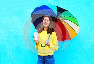 Happy pretty smiling young woman with coffee cup and colorful umbrella in autumn day over colorful blue background