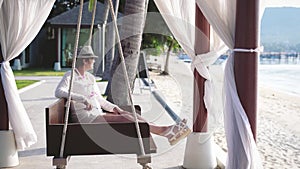 Happy pretty woman in aged in hat and sunglasses resting on a wooden swing on a tropical beach during summer vacation