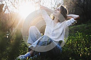 Happy pretty young cheerful woman in white shirt sitting on grass enjoying sunny spring day