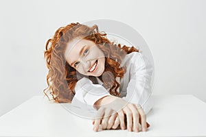 Happy pretty young girl with foxy hair smiling looking at camera sitting at table over white background.